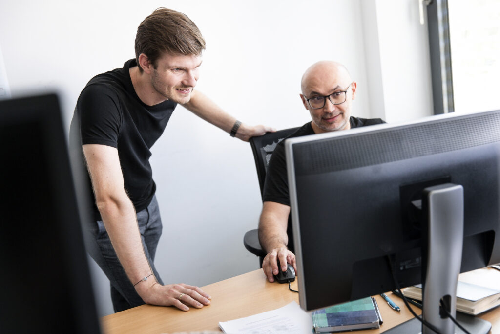 Zwei innovas-Mitarbeitende besprechen sich an einem PC an einem Arbeitsplatz.