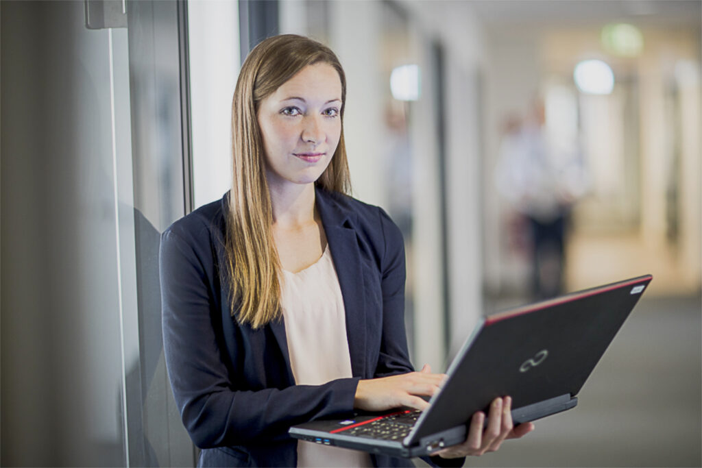 Eine innovas-Mitarbeitende steht mit ihrem Laptop vor ihrem Büro.