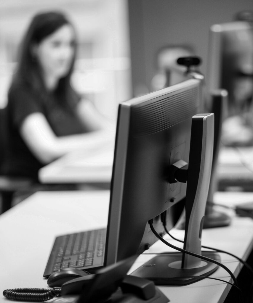 Im Vordergrund steht ein Monitor mit Telefon und im Hintergrund sitzt eine innovas-Mitarbeitende  an ihrem PC